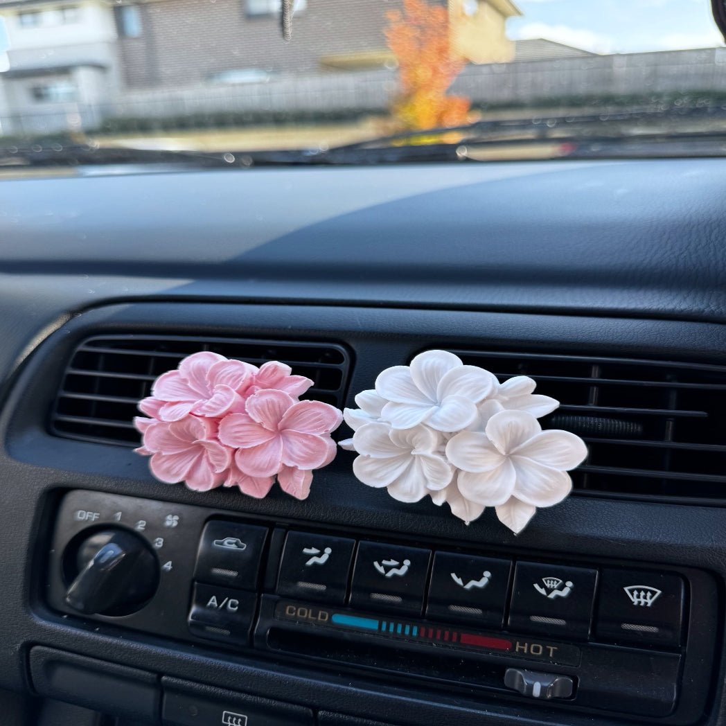Two Plumeria Eco Car Diffusers, one pink and one white, attached to a car's air vent.