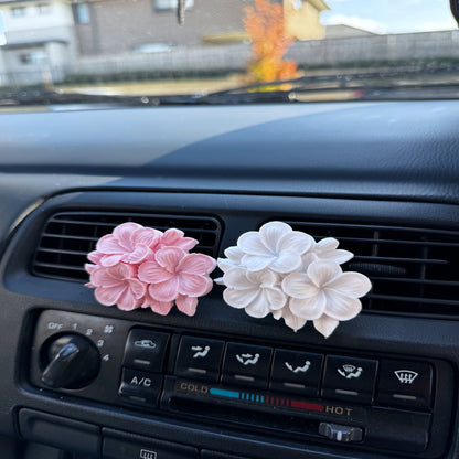 Two Plumeria Eco Car Diffusers, one pink and one white, attached to a car's air vent.